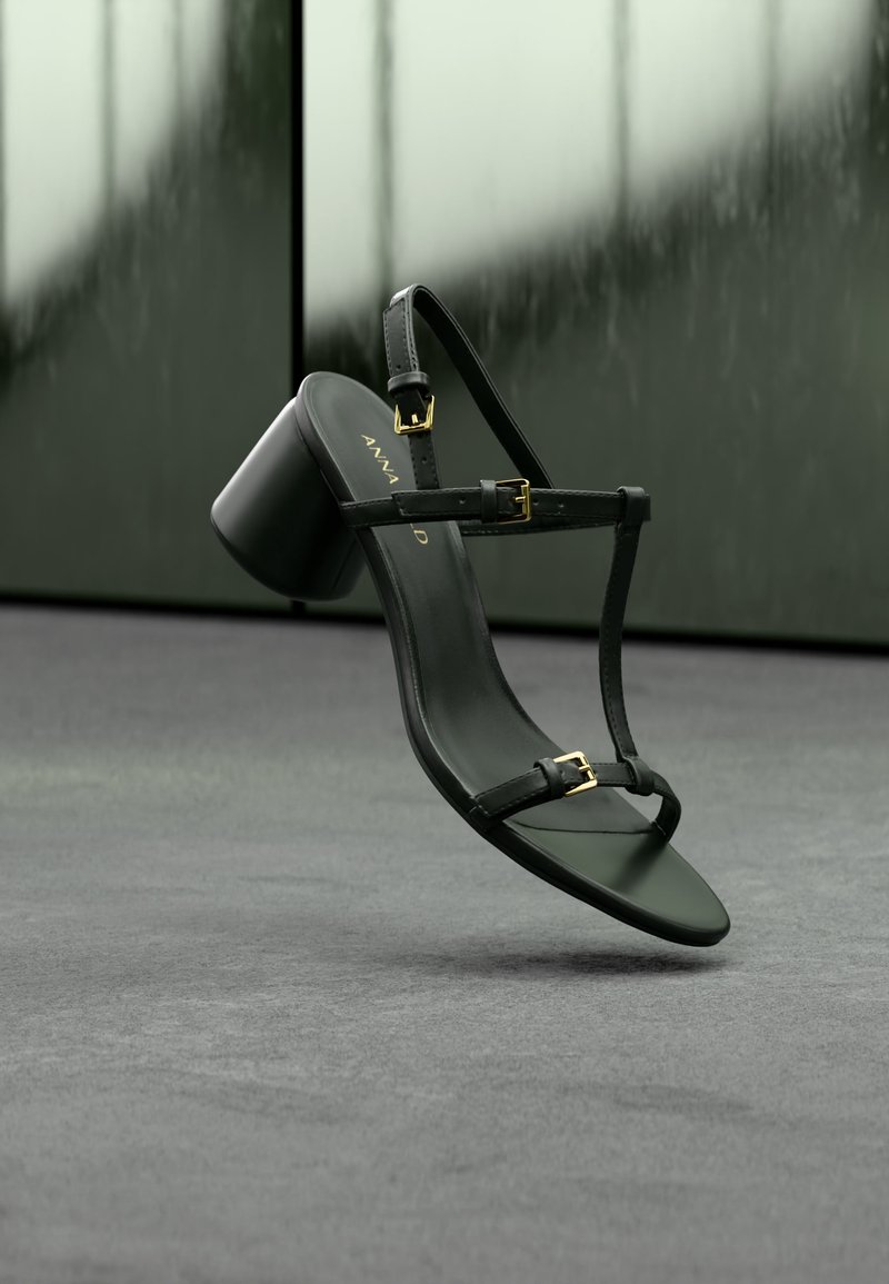 Μαύρο δερμάτινο σανδάλι με ψηλό τακούνι, λεπτές λουρίδες και χρυσά κουμπώματα, πάνω σε θολό φόντο με σκούρο πράσινο και γκρι αποχρώσεις.