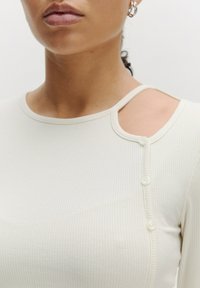 Licht beige geribde lange mouwen top met een uitsnede op de schouder, voorzien van drie ronde knopen aan de zijkant. Soepele textuur en aansluitend ontwerp.