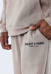Conjunto de sudadera y pantalones de color gris claro, hecho de tejido suave. Presenta el logo "PROJECT X PARIS MEMBERS' CLUB" en un estampado oscuro en los pantalones.