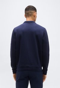 Άνδρας με την πλάτη, φοράει μπλε σκούρο πουλόβερ μακρυμάνικο με γιακά και αντίστοιχο μπλε παντελόνι, πάνω σε απλό φόντο.