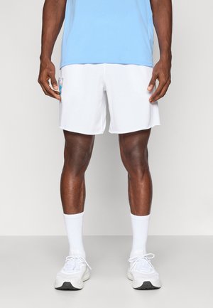 Homme portant un short de sport blanc, des chaussettes blanches, des baskets blanches et une chemise bleu clair, debout devant un fond blanc.