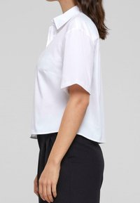 White short-sleeve shirt with a pointed collar, featuring a relaxed fit and a smooth texture, paired with dark trousers.
