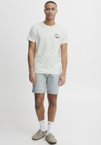 Lys beige t-shirt med et palmetræ grafik, parret med lyseblå shorts. Outfitten suppleres med grå slip-ons og hvide sokker.