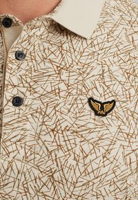 Polo a maniche corte beige con un motivo astratto marrone e verde. Presenta un colletto e un logo ricamato nero.