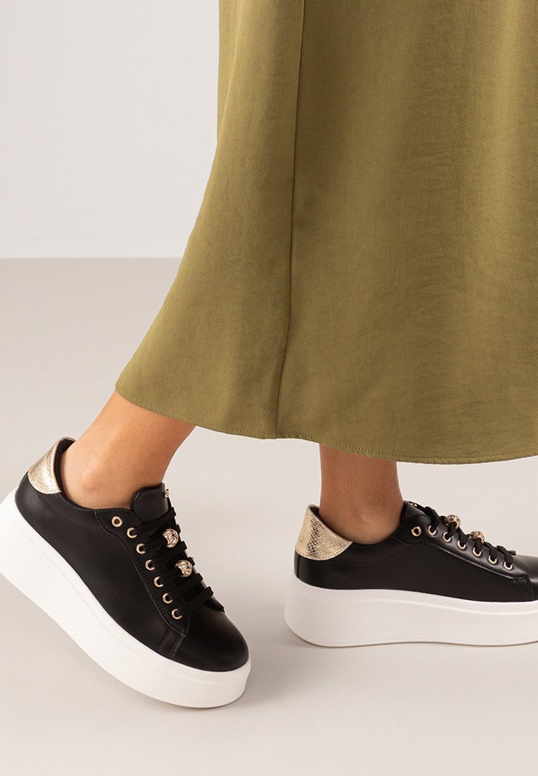 Zapatillas negras con suela de plataforma blanca, acentos metálicos dorados en el talón, ojales decorativos y parte superior de cuero negro texturizado.