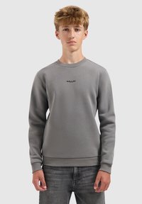 Jonge man die staat, gekleed in een grijze sweatshirt met het "BALLIN" logo centraal op de borst en vervaagde zwarte jeans, neutrale achtergrond.