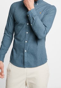 Blå skjorta med knappar och långa ärmar, tillverkad av texturerat tyg. Har en liten broderad logotyp och vita knappar.