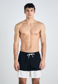 Svart og hvite svømmebukser med elastisk midje, snor som lukkemekanisme, og en kontrasterende horisontal hvit stripe nederst.
