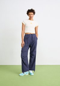 Top scurt alb cu textura ribbată, marginile galbene cu volane și un mic detaliu floral, asociat cu pantaloni largi în carouri bleumarin și adidași albaștri.