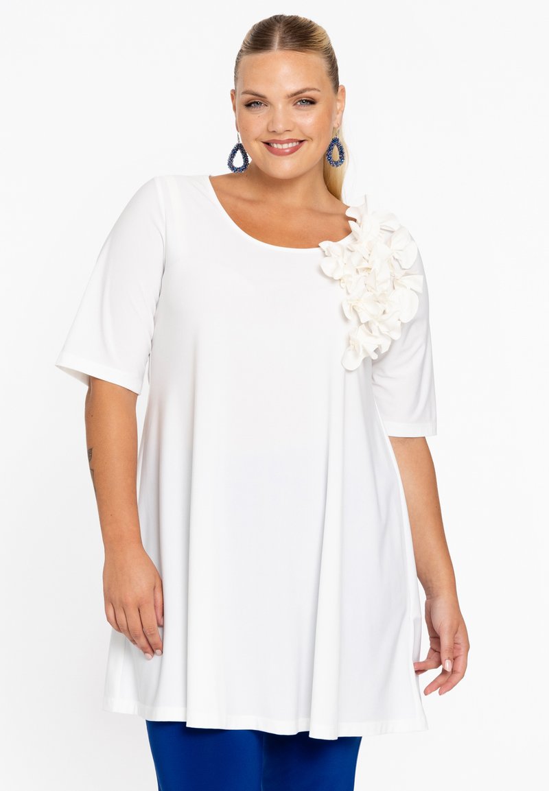 Haut tunique blanc à manches courtes, avec une accent floral texturé sur une épaule. Fabriqué en tissu doux ; design ample.