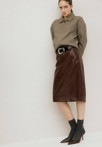 Kobieta w kurtce w jodełkę, brązowej skórzanej spódnicy midi z szerokim czarnym paskiem, szarymi skarpetkami i brązowych szpiczastych szpilkach, stojąca przy ścianie.