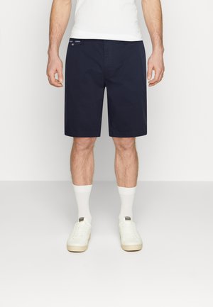 Homme portant un short bleu marine jusqu'aux genoux, des chaussettes blanches, des baskets blanches et un débardeur blanc, debout devant un fond uni.