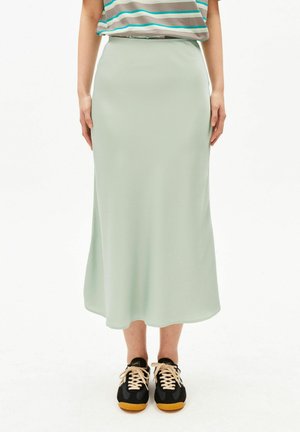 Frau trägt einen hellgrünen Midirock, ein gestreiftes Hemd und schwarze Sneakers mit beigen Schnürsenkeln, steht vor einem weißen Hintergrund.