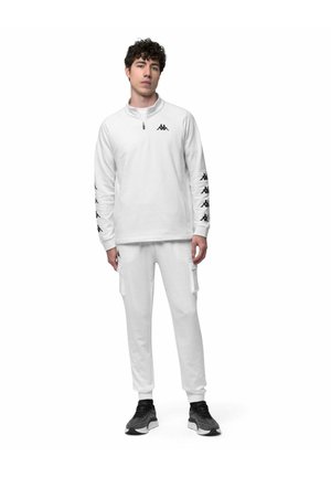 Giovane uomo con tuta bianca Kappa con logo sulle maniche e sul petto, abbinata a sneakers bianche e nere, in piedi davanti a uno sfondo bianco.