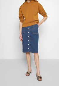 Denim skirt with a fitted silhouette, featuring a front button placket, two front pockets, and a raw hem. Paired with a brown hoodie and leopard print slide sandals.