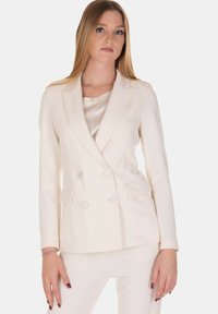 Blazer crema doppiopetto con una consistenza liscia, revers a lancia e due tasche frontali. Indossato sopra un top in seta abbinato.