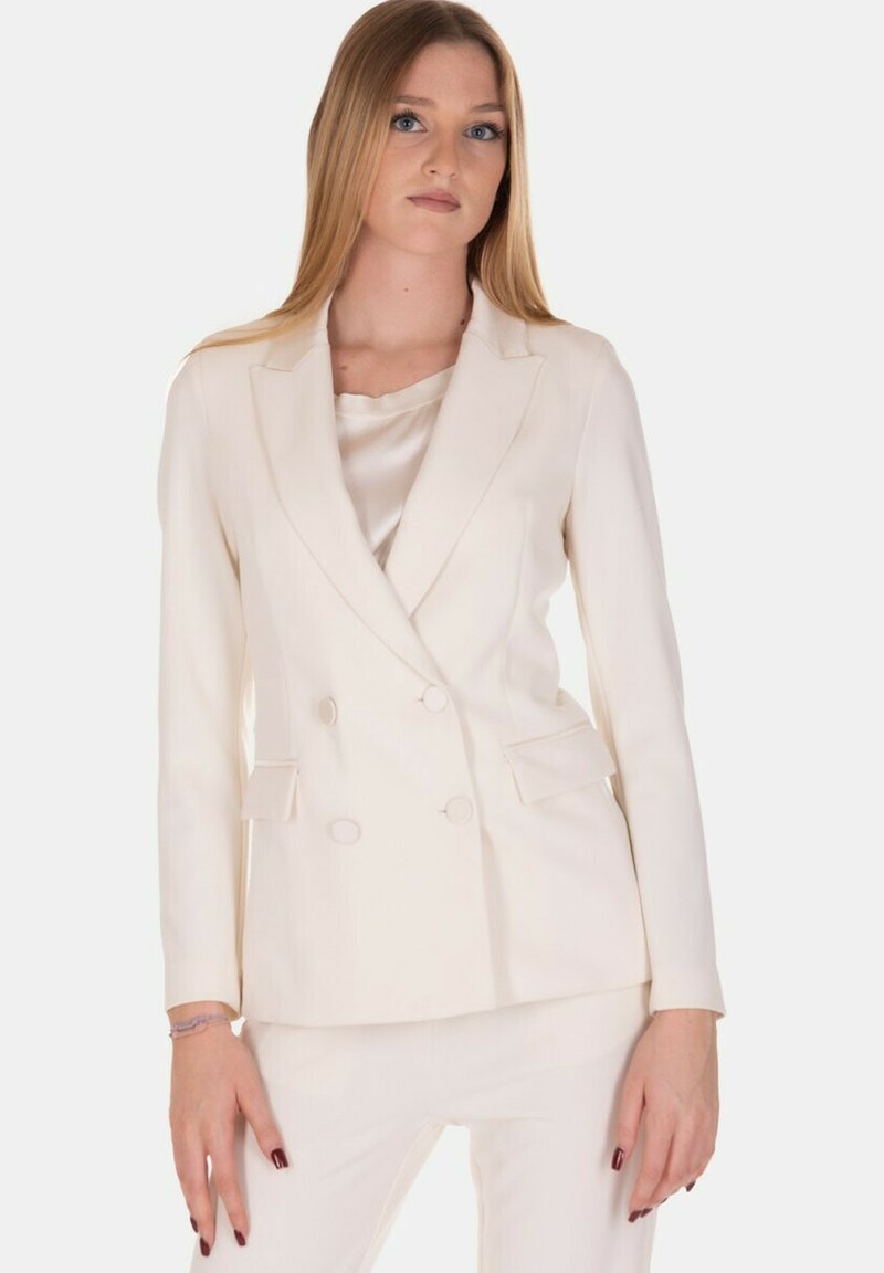 Blazer crema doppiopetto con una consistenza liscia, revers a lancia e due tasche frontali. Indossato sopra un top in seta abbinato.