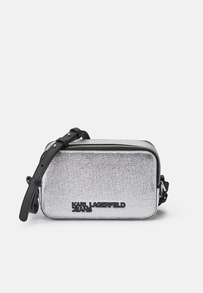 Sølvmetallisk crossbody-veske med glidelås, svart justerbar stropp og preget merkevare på fronten. Rektangulær form, glatt tekstur.