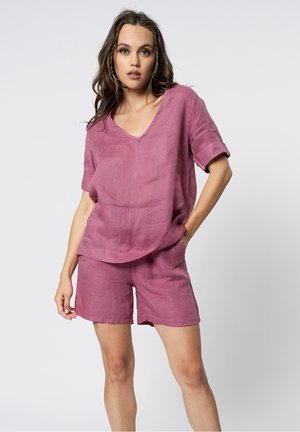 Donna in piedi con una mano in tasca, indossa una maglietta rosa a maniche corte e comoda e pantaloncini abbinati su uno sfondo semplice.