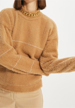 Vrouw die een pluizige lichtbruine trui met subtiele witte stiksels draagt, beige broek en een dikke gouden ketting, poserend met gebogen armen.