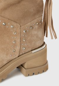 Bottines en suède beige avec des clous décoratifs en argent et en cristal, talon épais avec accent métallique et détail de franges sur le côté.