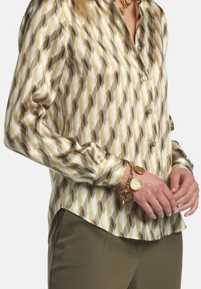 Blouse met lange mouwen van een satijnachtige stof met een geometrisch patroon in olijf-, crème- en bruine tinten; gecombineerd met een armband met gouden munten.