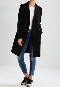 Musta kaksirivinen takki, jossa on kaulukset, yhdistettynä tiukan istuviin sinisiin farkkuihin ja mustiin lenkkareihin, joissa on vaaleanpunainen yksityiskohta. Sileä pinta.