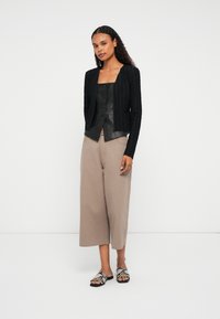 Kvinde med langt krøllet hår iført en sort struktureret cardigan over en sort knappet lædertop, beige brede forkortede bukser og sandaler i slangeskind.