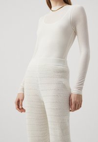 Bílý žebrovaný top s dlouhým rukávem v kombinaci s bílými širokými kalhotami s krajkovým vzorem, které mají texturovaný pletený materiál a elastický pas.