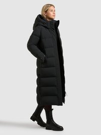 Manteau matelassé noir avec capuche, présentant un design long et matelassé avec des fermetures éclair sur les côtés. Porté avec des bottines noires et un pantalon foncé.