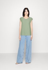 Haut vert à manches courtes avec des accents volants, associé à un jean bleu clair à jambes larges. La tenue présente un design décontracté et ample.