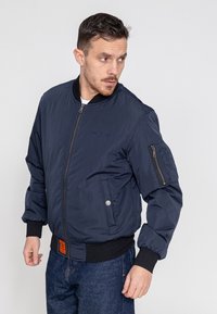 Marineblauwe bomberjack met een ritssluiting aan de voorkant, geribbelde manchetten en zijzakken. Bevat een geborduurd logo en een patch op de linker mouw.