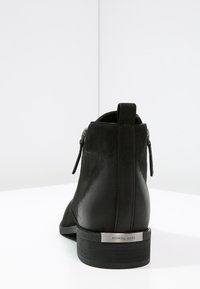 Bottines noires avec une combinaison de cuir lisse et texturé, présentant une fermeture éclair sur le côté et une plaque logo métallique argentée sur le talon.