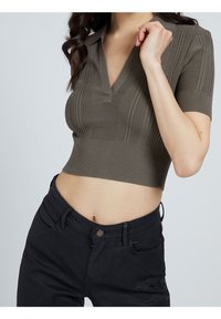 Haut en tricot court olive avec des manches courtes et un décolleté en V, présentant une texture en côtes et une patte discrète au niveau du col.