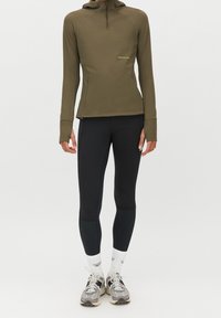 Pullover lungo a maniche con mezzi zip di colore verde oliva, abbinato a leggings neri. Le scarpe sono grigie con accenti neri e i calzini sono bianchi.