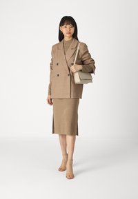 Blazer doppiopetto marrone indossato sopra un top in maglia beige e una gonna a matita beige lunghezza ginocchio. Sandali con cinturini trasparenti e una borsa leggera beige.