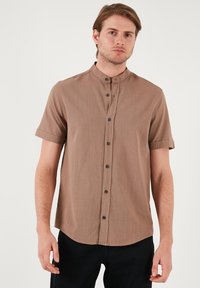 Korte mouwen bruine shirt met een mandarijnkraag, voorzien van zwarte knopen en een gestructureerde stof. Het shirt heeft een afgeronde onderkant en een ontspannen pasvorm.