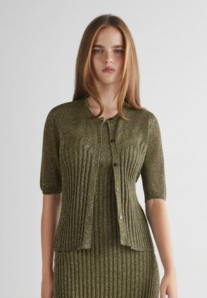 Giovane donna con capelli lisci castano chiaro che indossa un cardigan corto a coste verde oliva scintillante e una gonna abbinata.