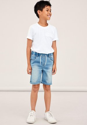 Valge lühikeste varrukatega t-särk, mis on kombineeritud helesiniste denim lühikeste pükstega, millel on paelaga vöökoht ja rullitud servad, kantud valgete spordijalgadega.