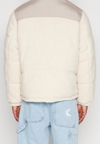 Krämliknande fleecejacka med en slät, ljusbeige nylonaxel. Har en boxig form och texturerad tyg. Kombineras med ljusblå denim.