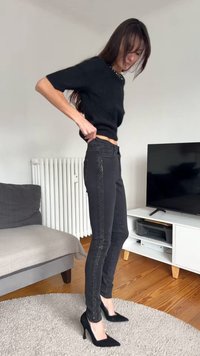 Sieviete tērpusies melnās pieguļošās džinsa biksēs ar sequined sānu svītrām un melniem augstpapēžu apaviem, regulē savas džinsas dzīvojamā istabā ar televizoru un dīvānu.