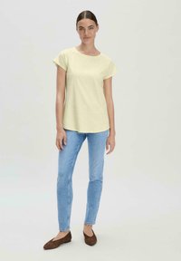 Lichtgele, kortemouwen t-shirt met een ronde hals, gecombineerd met blauwe jeans en bruine flats, staand tegen een neutrale achtergrond.