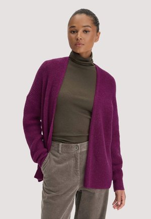 Frau mit zurückgebundenen Haaren, die einen lila offenen Cardigan, einen olivgrünen Rollkragenpullover und beige Cordhosen trägt, steht vor einem einfarbigen Hintergrund.