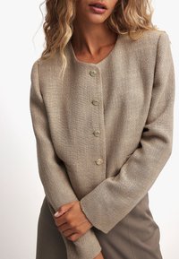 Beige texturerad jacka med rund halsringning, utrustad med fem framknappar och långa ärmar, parat med en figursydd grå kjol.