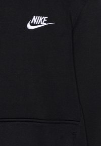 Tela negra con el logo bordado en blanco de Nike y el swoosh en la parte superior izquierda, costuras visibles y una costura de bolsillo debajo del logo.