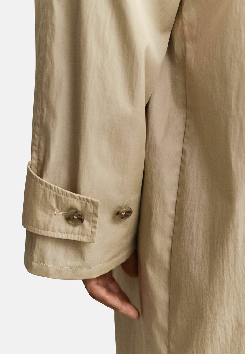 Nahaufnahme eines beigen Trenchcoat-Ärmels mit zwei Knöpfen an der Manschette und einer Hand, die neben dem Mantel ruht.