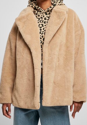 Beige faux fur coat worn over a beige and black spotted hoodie and blue jeans, shown from shoulders to hips.