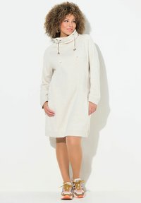 Helles beige Kleid mit Kapuze aus weichem Stoff, mit langen Ärmeln und Zugbändern. Getragen mit mehrfarbigen Turnschuhen.