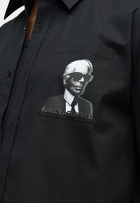 Μαύρο πουκάμισο με τσέπη στο στήθος που έχει ασημένιο γραφικό ενός ατόμου με γυαλιά ηλίου και κείμενο "KARL LAGERFELD" από κάτω.