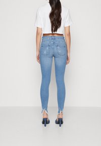 Mujer con cabello largo y oscuro, vistiendo unos ajustados vaqueros desgastados de color azul claro, una camiseta blanca corta y zapatos de tacón azules, de pie de espaldas.